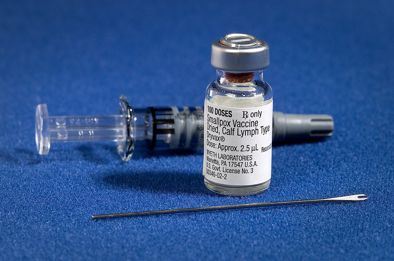 A vial of smallpox vaccine shown alongside a bifurcated needle and syringe.